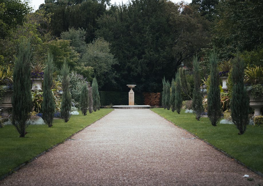 A symmetrical garden path with a gravel surface extends straight ahead, flanked by neatly trimmed grass lawns and planted borders containing various lush plants and shrubs. Tall, slender cypress trees